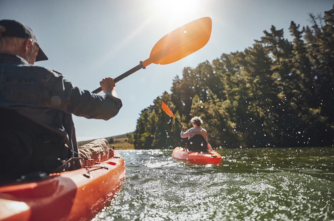 Outdoor shot of senior man canoeing in the lake with woman in background on a summer day. Man and woman in two different kayaks in the lake on a sunny day